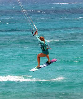 Une kitesurfeuse s'élance hors de l'eau sur une planche, tirée par une aile, sur les eaux turquoise de Saint-Pierre-la-Mer, sous un ciel ensoleillé.