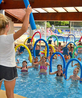 Een groep kinderen en een volwassene die deelnemen aan een wateractiviteit in een overdekt zwembad, waarbij ze gebruikmaken van zwemnoodles en kickboards, met op de achtergrond een glijbaan zichtbaar.